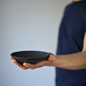 May include: A person holds out a black, round, ceramic bowl with a slightly flared rim.