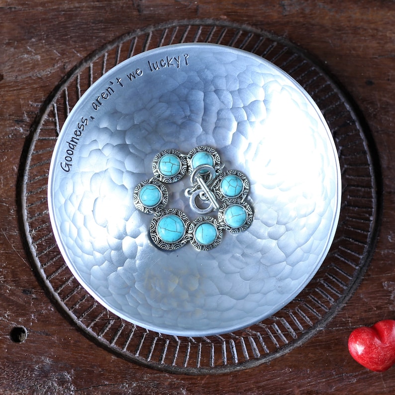May include: A round, hammered silver-tone metal bowl with the text "Goodness, aren't we lucky?" stamped along the inner rim. Inside the bowl sits a bracelet featuring circular silver-tone links with turquoise-coloured stone inlays and a toggle clasp.