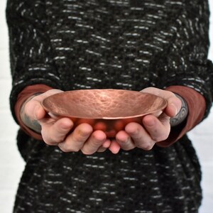 May include: A hammered copper bowl with a slightly rounded bottom. The bowl is held in the hands of a person.