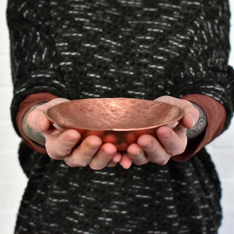 May include: A hammered copper bowl with a slightly rounded bottom. The bowl is held in the hands of a person.