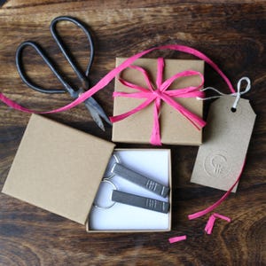 May include: Two silver keychains with a textured design are displayed in a small, white box. The box is open and sitting on a wooden surface. A brown gift box with a pink ribbon is in the background.