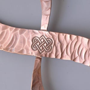 May include: Close-up of a copper-colored metal object with a textured surface and a Celtic knot design in the center. The metal has a hammered appearance, and the knot is a lighter silver color.