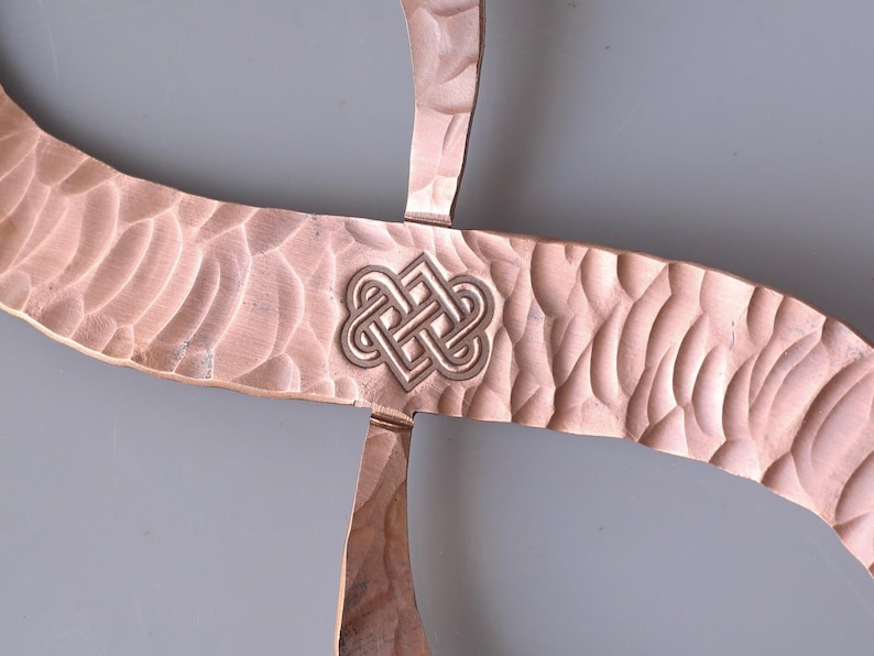 May include: Close-up of a copper-colored metal object with a textured surface and a Celtic knot design in the center. The metal has a hammered appearance, and the knot is a lighter silver color.