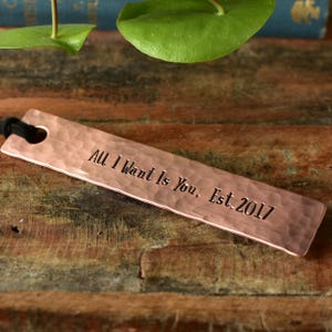 May include: Copper bookmark with the text "All I Want Is You, Est. 2017." The bookmark has a hammered texture and a black cord. The bookmark is rectangular with a heart-shaped hole at the top.