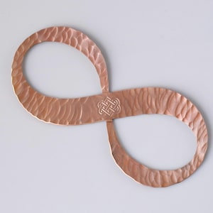 May include: A copper-colored infinity symbol with a textured surface and a Celtic knot design in the center. The metal art piece is displayed against a white background. The infinity symbol is a decorative item.