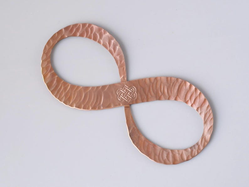 May include: A copper-colored infinity symbol with a textured surface and a Celtic knot design in the center. The metal art piece is displayed against a white background. The infinity symbol is a decorative item.