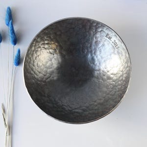 May include: A dark grey, circular metal bowl with a hammered, dimpled texture on its interior surface. The rim of the bowl features the engraved text "Est. 2015" in a serif font. The bowl is positioned next to four thin, blue, dried floral stems.