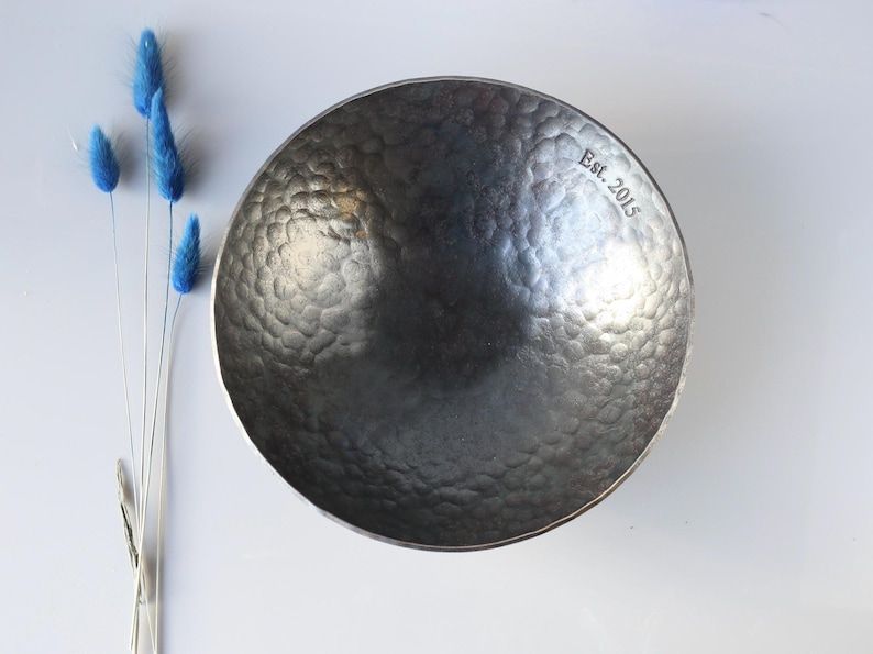 May include: A dark grey, circular metal bowl with a hammered, dimpled texture on its interior surface. The rim of the bowl features the engraved text "Est. 2015" in a serif font. The bowl is positioned next to four thin, blue, dried floral stems.
