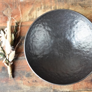 May include: A dark grey, circular, hammered metal bowl with a textured surface. The rim is engraved with the text "David & Elaine 01 May 2024". Beside the bowl is a small, tied bundle of dried beige grasses and green eucalyptus leaves.