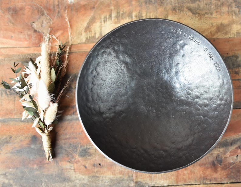 May include: A dark grey, circular, hammered metal bowl with a textured surface. The rim is engraved with the text "David & Elaine 01 May 2024". Beside the bowl is a small, tied bundle of dried beige grasses and green eucalyptus leaves.