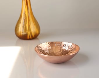 May include: A small, round, hammered copper bowl with a metallic finish. The interior rim is stamped with the text L heart J 22.06.19. The bowl has a textured, dimpled surface and a warm, reddish-brown color, reflecting light on its curved exterior.