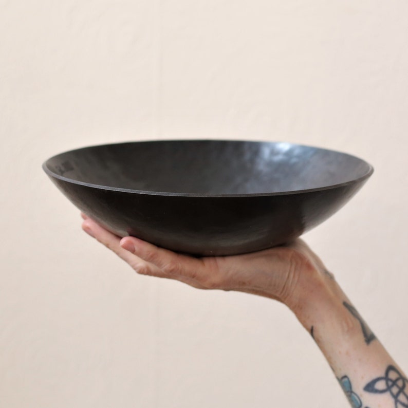 May include: A shallow, circular bowl with a dark, matte finish and a subtly hammered, textured surface. The bowl features a wide, open rim and a smooth, curved interior, showcasing a consistent dark charcoal or black colour throughout the exterior.