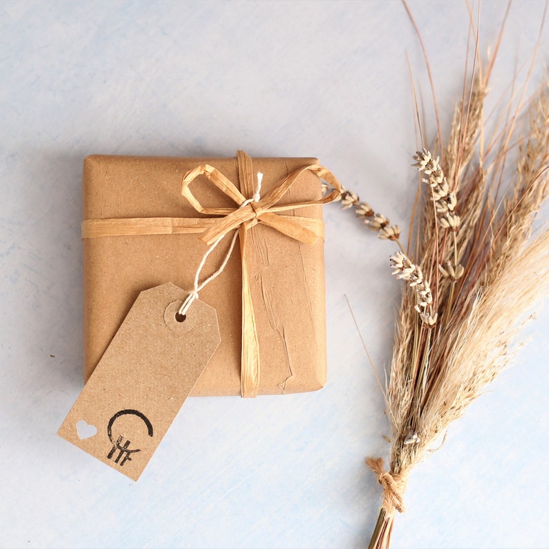 Peut inclure: Une bo&icirc;te cadeau carr&eacute;e emball&eacute;e dans du papier kraft brun, nou&eacute;e avec un ruban en raphia beige. Une &eacute;tiquette rectangulaire en papier brun est attach&eacute;e au ruban, avec une petite ic&ocirc;ne de c&oelig;ur et un logo circulaire noir &agrave; trois lignes.