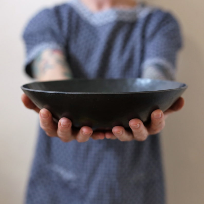 May include: A shallow, circular bowl with a matte black finish and a smooth, curved interior. The bowl features a wide, open rim and a consistent dark colour throughout its surface, presenting a simple, functional design for serving or display.