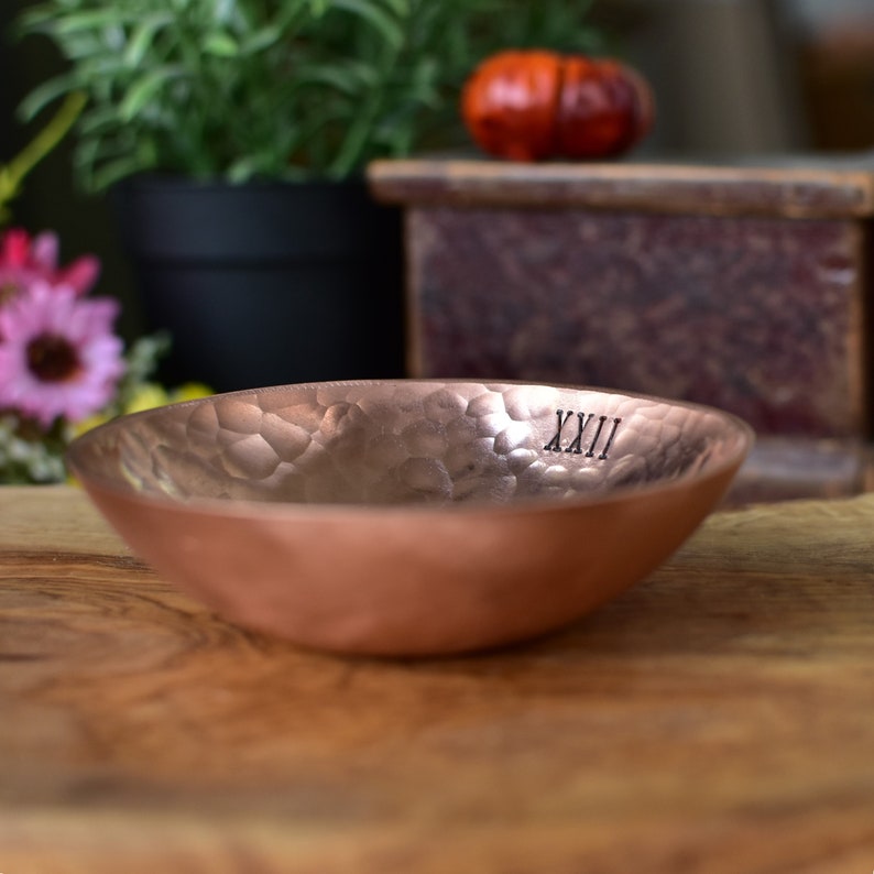 Peut inclure: Un bol circulaire peu profond en cuivre pr&eacute;sentant une texture martel&eacute;e distincte sur sa surface int&eacute;rieure. Le m&eacute;tal poss&egrave;de une finition m&eacute;tallique brun rouge&acirc;tre chaleureuse. Les chiffres romains XXII sont imprim&eacute;s en noir sur le bord.