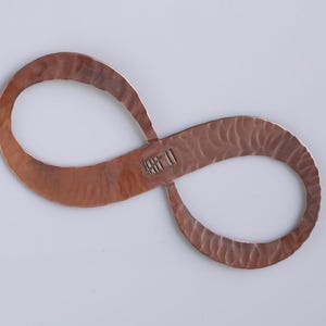 May include: A copper-colored infinity symbol with a textured surface. The metal has a hammered appearance, and the symbol has a small, stamped marking in the center. The shape is displayed against a plain, light background.