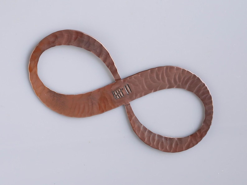 May include: A copper-colored infinity symbol with a textured surface. The metal has a hammered appearance, and the symbol has a small, stamped marking in the center. The shape is displayed against a plain, light background.