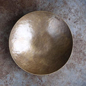 May include: A round, hammered brass bowl with a metallic gold finish. The interior surface features a textured, dimpled pattern throughout. The word "NOA" is stamped in a simple, dark font near the upper right rim of the bowl's interior.