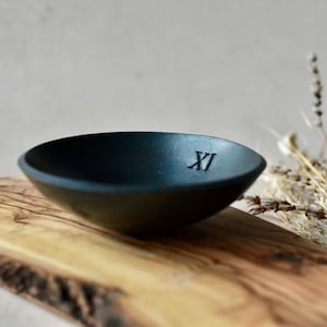 May include: A small, black, round bowl with a Roman numeral 'XI' engraved on the bottom. The bowl is sitting on a wooden surface.