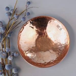 May include: A hammered copper bowl with a smooth, shiny finish. The bowl has a slightly rounded shape and a small inscription on the bottom that reads "HT II".