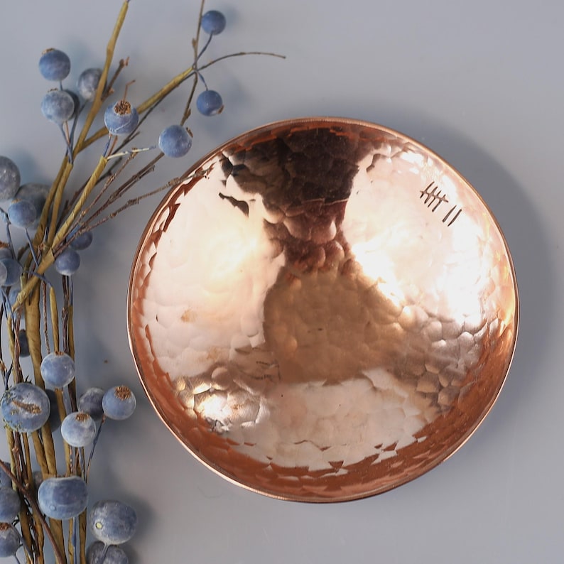 May include: A hammered copper bowl with a smooth, shiny finish. The bowl has a slightly rounded shape and a small inscription on the bottom that reads "HT II".