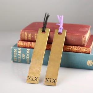 May include: Two rectangular brass bookmarks with a smooth finish, each featuring the Roman numeral XIX stamped at the bottom. One bookmark has a black cord tied through the top hole, while the other has a light purple cord tied through its top hole.