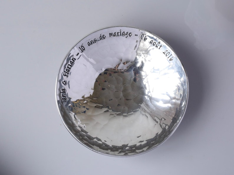May include: A round, silver-coloured bowl with a hammered, reflective surface. The inner rim features black engraved text that reads: Tania & Florian - 10 ans de mariage - 06 ao&ucirc;t 2016. The bowl has a smooth, polished finish and a circular shape.