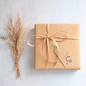 May include: A square gift box wrapped in plain brown paper, tied with a thin raffia ribbon bow. A small rectangular brown paper tag is attached to the ribbon, featuring a black stamped logo with a circle, lines, and a small heart symbol.