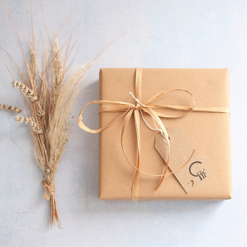May include: A square gift box wrapped in plain brown paper, tied with a thin raffia ribbon bow. A small rectangular brown paper tag is attached to the ribbon, featuring a black stamped logo with a circle, lines, and a small heart symbol.