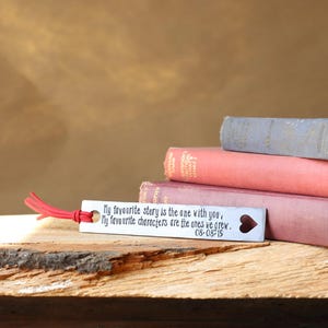 Può includere: Un segnalibro rettangolare in metallo color argento con una nappa in corda rossa. Il testo recita: My favourite story is the one with you, My favourite characters are the ones we grew. 08-08-15. A destra è presente un intaglio a cuore.