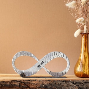 May include: A silver-colored infinity symbol sculpture with a textured surface and black tally marks in the center. The sculpture is displayed on a wooden surface next to a brown glass vase with dried flowers.