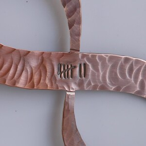 May include: Close-up of a copper-colored metal sculpture with a textured, hammered finish. The design is a cross-like shape with curved arms. The center features etched markings. The sculpture is likely a decorative wall hanging.
