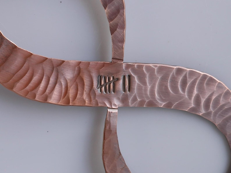 May include: Close-up of a copper-colored metal sculpture with a textured, hammered finish. The design is a cross-like shape with curved arms. The center features etched markings. The sculpture is likely a decorative wall hanging.