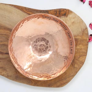 May include: A round, hammered copper bowl with a polished metallic finish. The interior rim features the engraved text "Every Year Better Than The Last" in a simple, dark font. The bowl has a textured, dimpled surface throughout its interior.