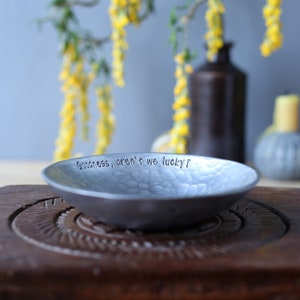 May include: A shallow, circular metal bowl with a hammered, textured finish. The interior rim is stamped with the black text "Goodness, aren't we lucky?". The bowl has a matte grey appearance and a slightly irregular, organic edge profile.