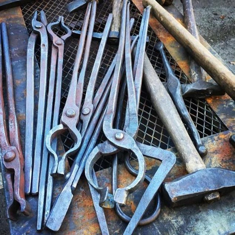 May include: A collection of dark grey forged steel blacksmithing tools including several pairs of long-handled tongs with varied jaw shapes and a heavy metal hammer with a wooden handle, all arranged on a metal grid surface.