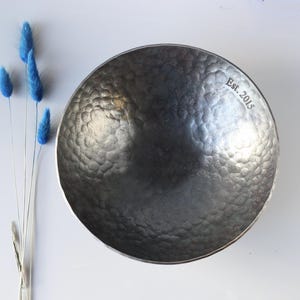 May include: A dark grey, circular metal bowl with a hammered, textured surface. The interior rim features the engraved text Est. 2015. The bowl has a deep, rounded basin and a metallic finish that reflects light across its uneven, dimpled surface.
