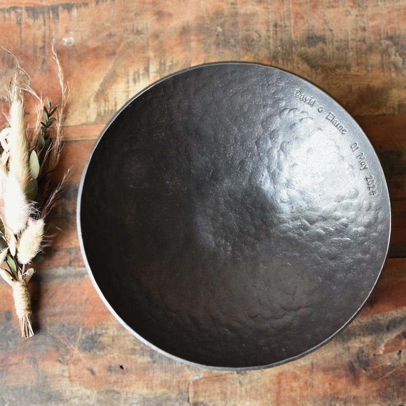 May include: A round, dark grey, hammered metal bowl with a textured surface. The rim is engraved with the text David & Elaine 01 May 2024. Beside the bowl is a small decorative bundle of dried grasses and green leaves tied with twine.