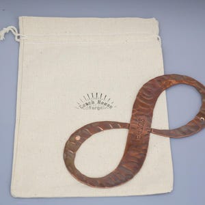 May include: A copper-colored metal infinity symbol with a textured surface, resting on a cream-colored canvas bag. The symbol has the text "Est. 2018" and is likely a decorative item or a piece of art.