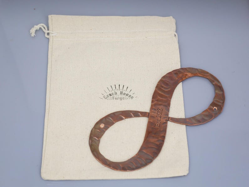 May include: A copper-colored metal infinity symbol with a textured surface, resting on a cream-colored canvas bag. The symbol has the text "Est. 2018" and is likely a decorative item or a piece of art.