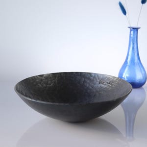 May include: A dark grey, shallow, circular bowl with a hammered, textured surface. The interior rim of the bowl features the engraved text "Est. 2021". The bowl has a matte finish and a wide, open design, presented as a standalone functional object.