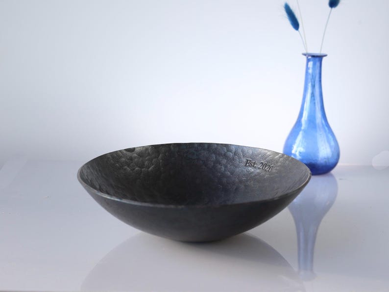 May include: A dark grey, shallow, circular bowl with a hammered, textured surface. The interior rim of the bowl features the engraved text "Est. 2021". The bowl has a matte finish and a wide, open design, presented as a standalone functional object.