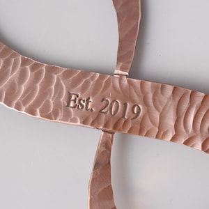 May include: Copper-colored metal art piece with a hammered texture. The design features curved, intersecting lines. The text "Est. 2019" is engraved on the central bar. The piece has a warm, metallic finish.