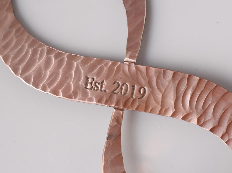 May include: Copper-colored metal art piece with a hammered texture. The design features curved, intersecting lines. The text "Est. 2019" is engraved on the central bar. The piece has a warm, metallic finish.
