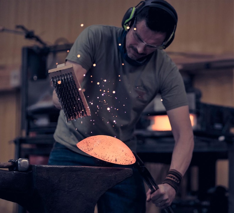 May include: A blacksmith working in a workshop, using a brush on a glowing, orange-hot metal object. Sparks fly as the artisan works, wearing safety glasses and ear protection. The anvil is visible in the foreground.