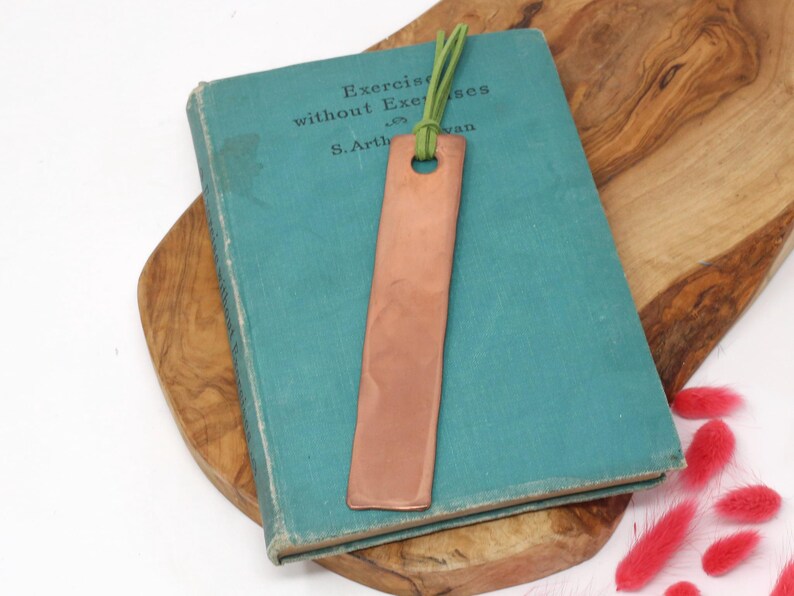 Pu&ograve; includere: Un segnalibro in rame con un cordoncino in pelle verde. Il segnalibro &egrave; appoggiato su un libro blu-verde con il testo "Exercise without Exercises" sulla copertina.