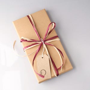 May include: A rectangular gift box wrapped in brown paper, tied with burgundy and cream-colored ribbons. A small, light brown tag hangs from the ribbon, with a simple design. The overall presentation is neat and elegant.