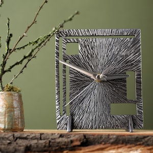 May include: A silver metal clock with a textured face and a geometric design. The clock has a silver metal frame and hands.