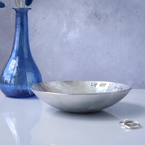 May include: A shallow, hammered silver-toned metal bowl with a polished finish. The inner rim is engraved with two sets of tally marks and the date 07-16-2016. The bowl has a curved, smooth exterior and a wide, open circular opening.