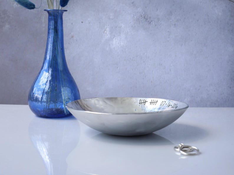 May include: A shallow, hammered silver-toned metal bowl with a polished finish. The inner rim is engraved with two sets of tally marks and the date 07-16-2016. The bowl has a curved, smooth exterior and a wide, open circular opening.